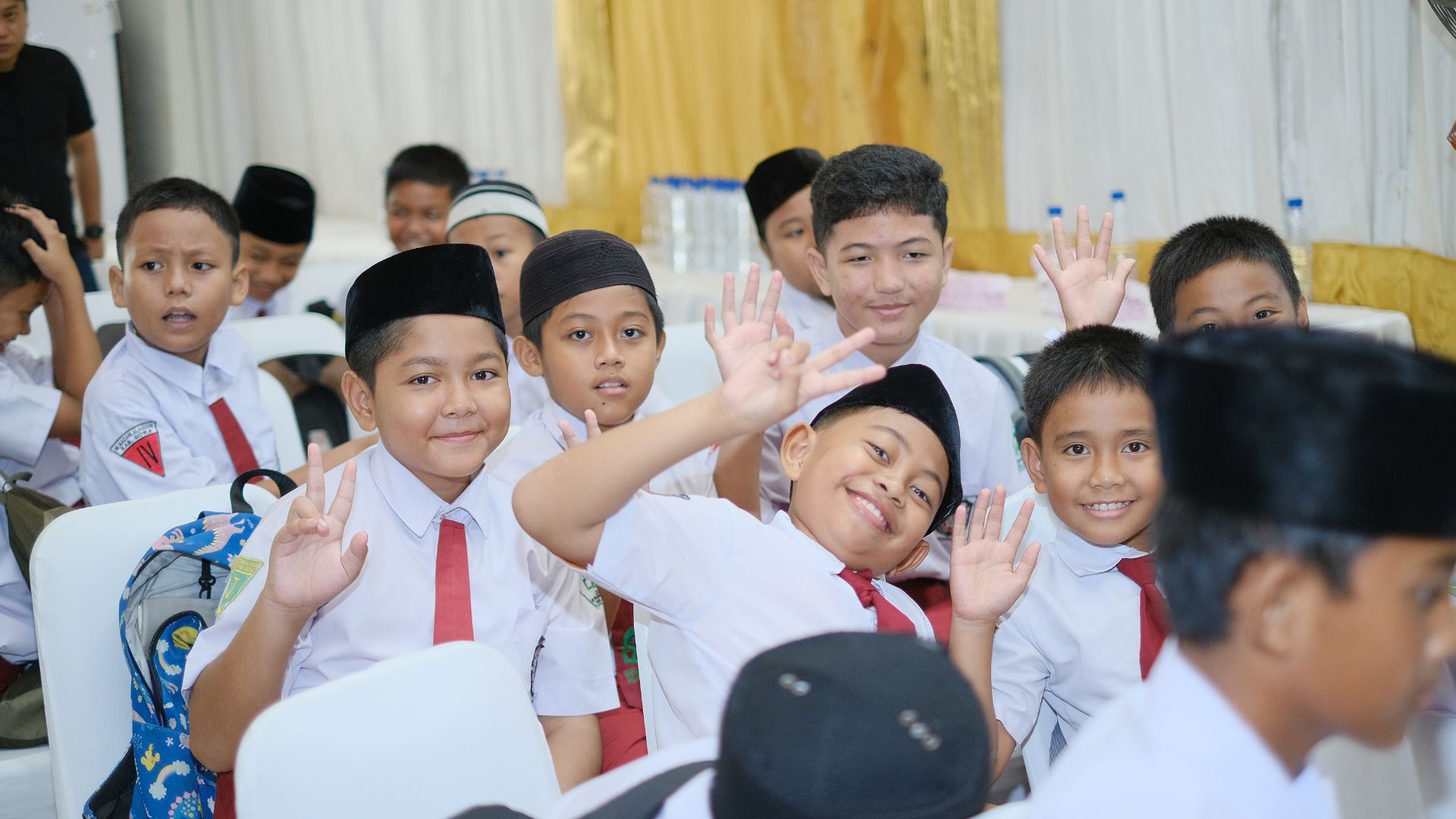 Anak-anak laki-laki yang tersenyum mengenakan seragam dan topi, berpose dan melambaikan tangan.