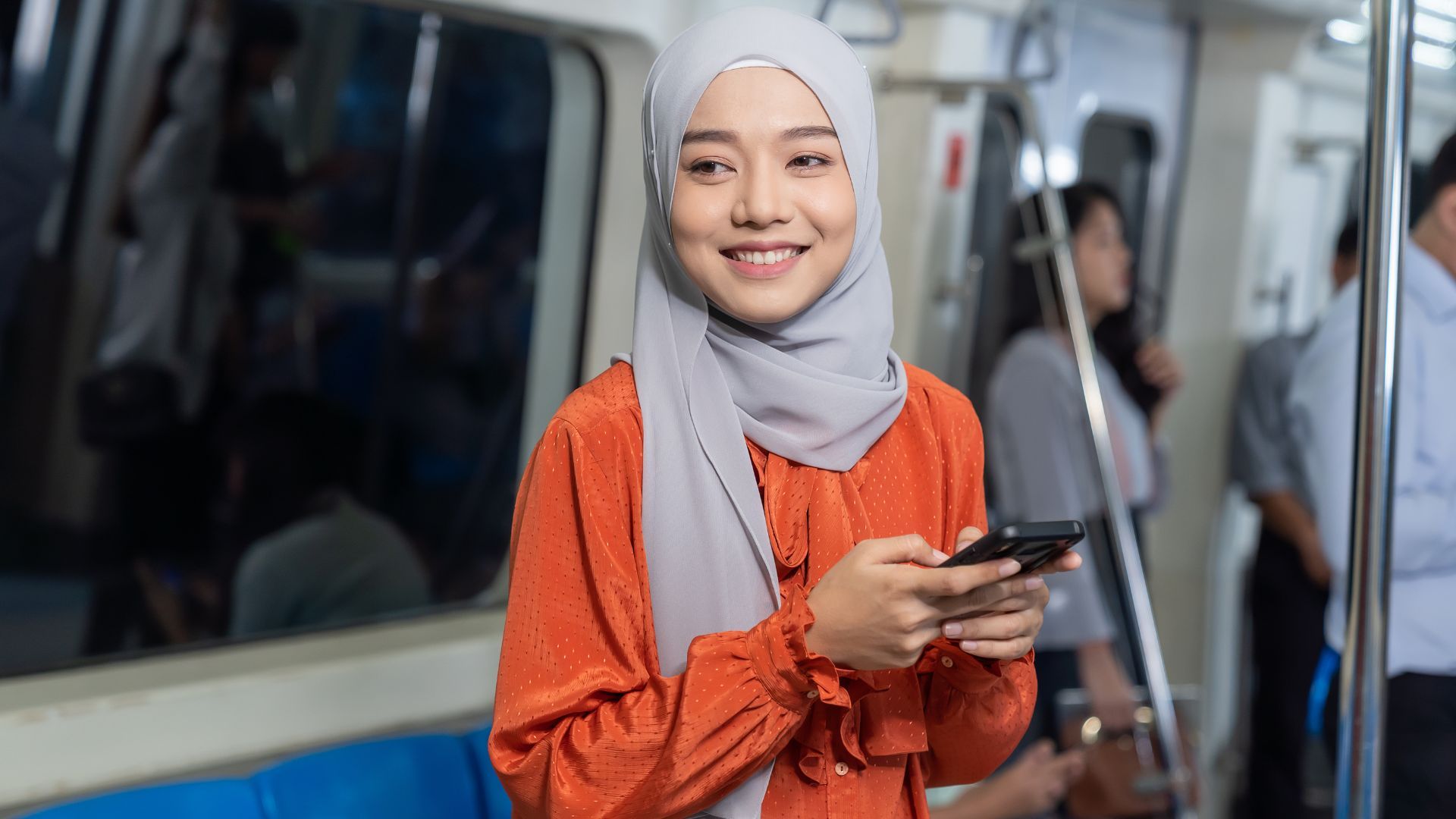 Seorang wanita berbaju blus oranye tersenyum dan memegang telepon di dalam kereta.