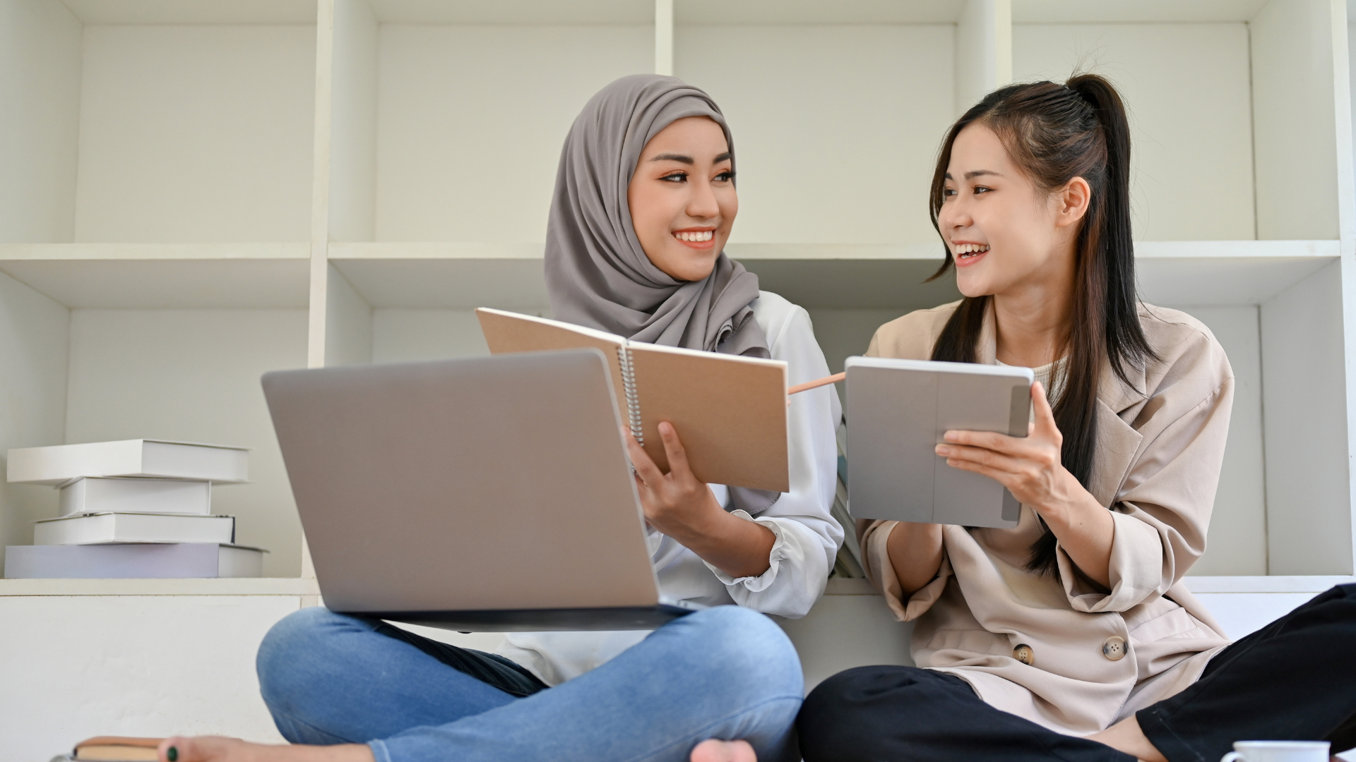 Dua wanita tersenyum sambil belajar menggunakan laptop dan buku catatan.